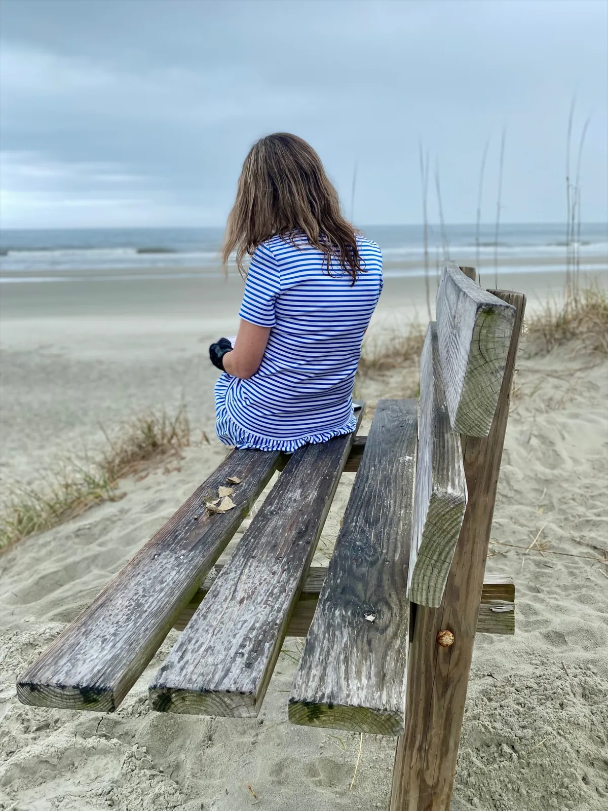 Une femme de dos, poratnt un maillot bleu et blanc rayé, assise sur un banc, regarde l'horizon du coté del'océan.